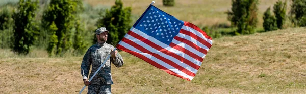 Tiro panorâmico de homem militar bonito em uniforme e cap segurando bandeira americana — Fotografia de Stock
