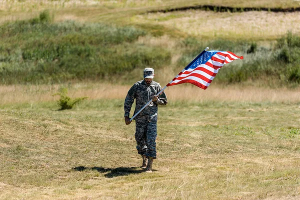 Handsome military man in uniform and cap walking and holding american flag in summertime — Stock Photo