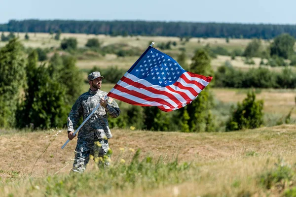 Selective focus of handsome military man in uniform and cap holding american flag in summertime — Stock Photo