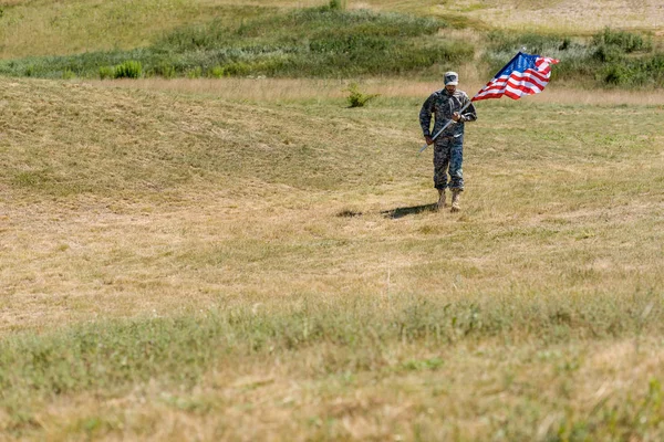 Selective focus of handsome soldier in uniform walking and holding american flag in summertime — Stock Photo