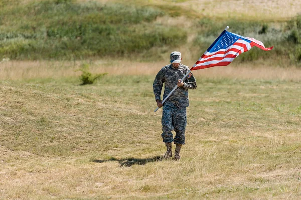 Foco seletivo do soldado em uniforme andando e segurando bandeira americana no verão — Fotografia de Stock