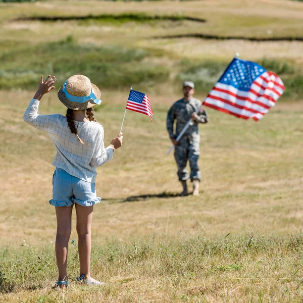 Back view of child in straw hat waving hand while holding american flag near father in military uniform — Stock Photo