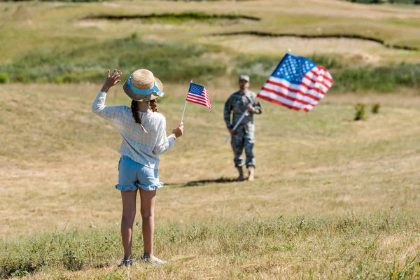 Vista trasera del niño en sombrero de paja agitando la mano mientras sostiene la bandera americana cerca del padre en uniforme militar - foto de stock