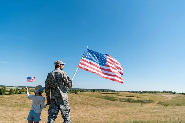 Soldier holding hands with kid in straw hat and holding american flag — Stock Photo