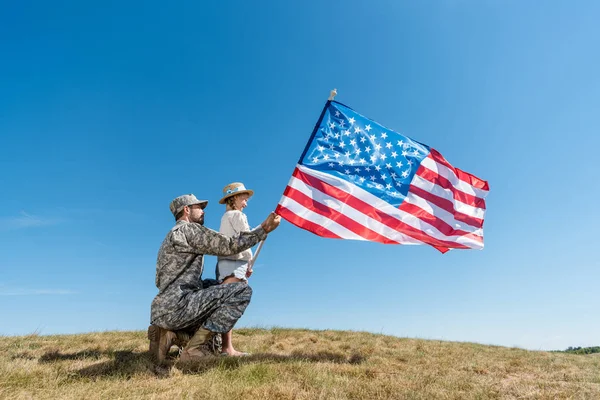 Pai em uniforme militar e criança segurando bandeira americana contra o céu — Fotografia de Stock