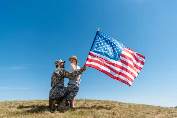 Father in military uniform and happy kid holding american flag against sky — Stock Photo