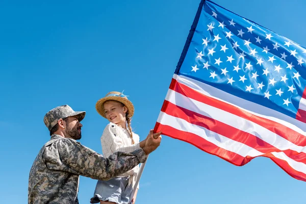 Low angle view of father in military uniform and happy kid holding american flag against sky — Stock Photo