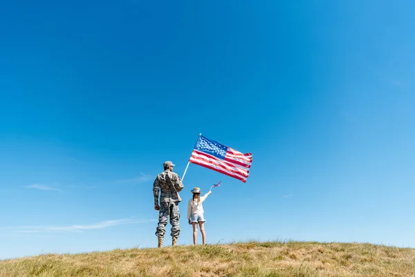 Vue arrière du gamin en chapeau de paille et père militaire tenant des drapeaux américains — Photo de stock