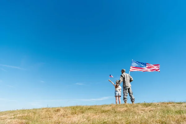 Vue arrière du gamin en chapeau de paille et père militaire tenant des drapeaux américains tout en se tenant debout sur l'herbe — Stock Photo