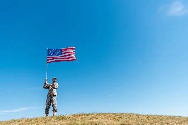 Vista ad angolo basso di bel militare in uniforme che cammina con bandiera americana con stelle e strisce — Foto stock