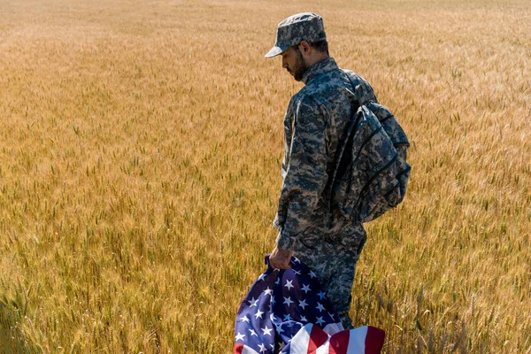 Патриотический солдат в военной форме, держащий американский флаг, стоя в поле с пшеницей — стоковое фото