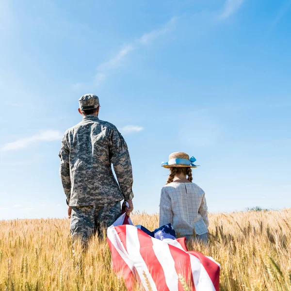 Visão traseira de soldado e criança em pé no campo e segurando bandeira americana — Fotografia de Stock