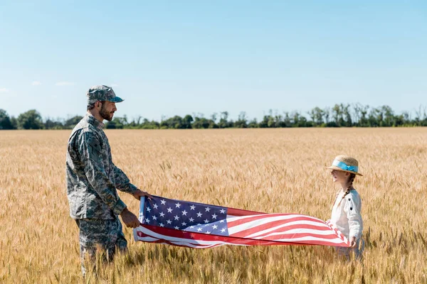 Soldado en uniforme y niño alegre sosteniendo bandera americana en el campo - foto de stock