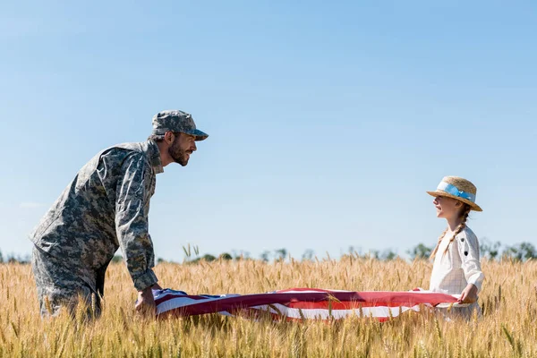 Soldier in uniform and happy child holding american flag in field — Stock Photo