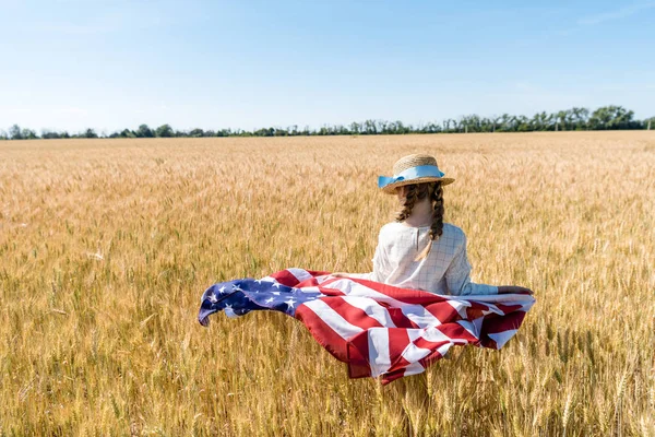 Vue arrière du gamin en chapeau de paille tenant le drapeau américain dans le champ d'or en été — Photo de stock