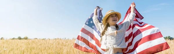 Panoramic shot of happy child in straw hat holding american flag in golden field with wheat — Stock Photo