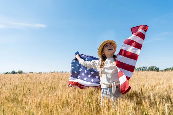 Cheerful child in straw hat holding american flag in golden field with wheat — Stock Photo