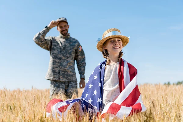 Избирательный фокус веселого ребенка, стоящего с американским флагом рядом с солдатом — стоковое фото
