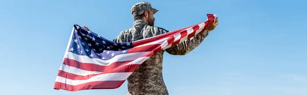 Panoramic shot of soldier in cap and uniform holding american flag against blue sky — Stock Photo