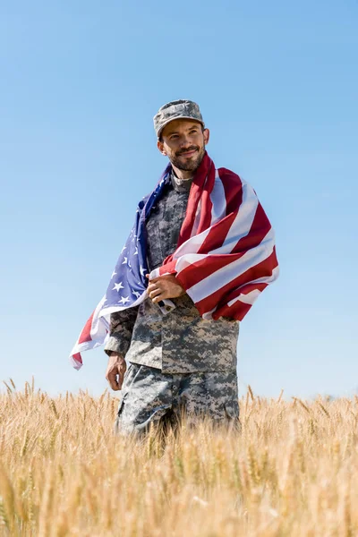 Selective focus of happy soldier in cap and uniform holding american flag in field — Stock Photo