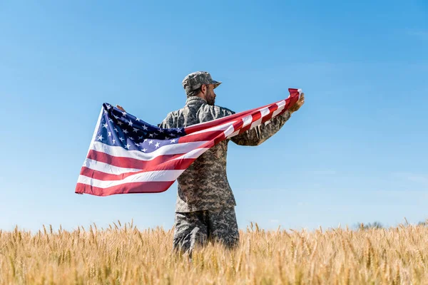 Soldier in cap and uniform holding american flag in golden field with wheat — Stock Photo