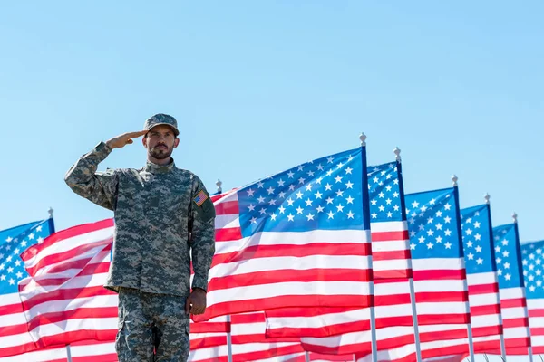 Mann in Militäruniform salutiert in der Nähe amerikanischer Flaggen mit Sternen und Streifen — Stockfoto