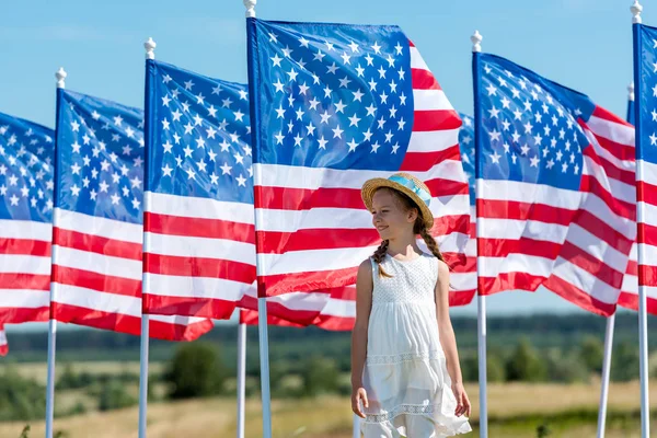 Niedliches patriotisches Kind, das im weißen Kleid neben amerikanischen Flaggen steht — Stockfoto