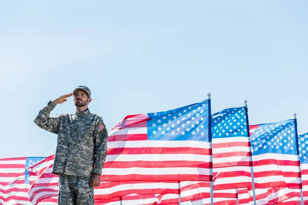 Soldado patriótico en uniforme militar dando saludo cerca de banderas americanas con estrellas y rayas — Stock Photo