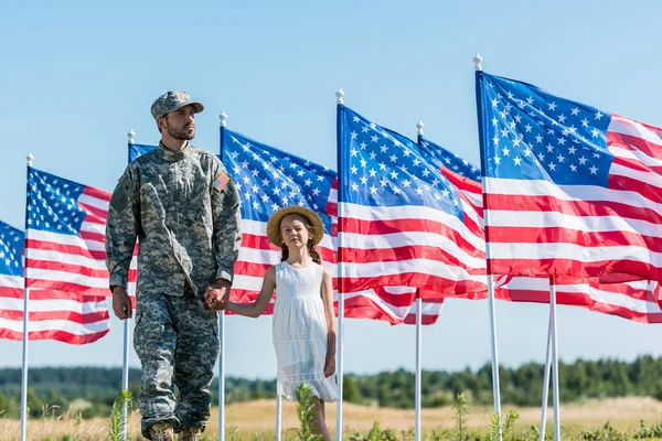 Красивый солдат, держащийся за руки с симпатичным ребенком возле американских флагов — стоковое фото