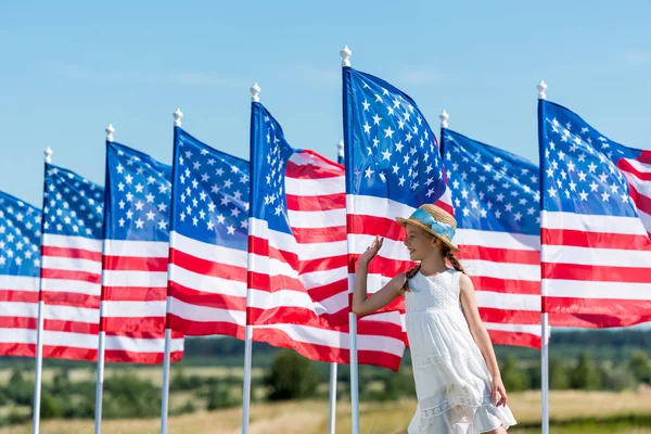 Glückliches patriotisches Kind, das in weißem Kleid neben amerikanischen Fahnen steht und Hand schwenkt — Stockfoto