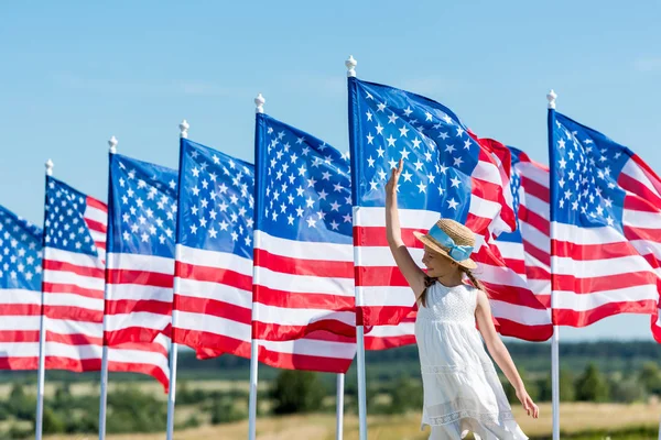 Fröhliches patriotisches Kind steht in weißem Kleid neben amerikanischen Fahnen und schwenkt die Hand — Stockfoto