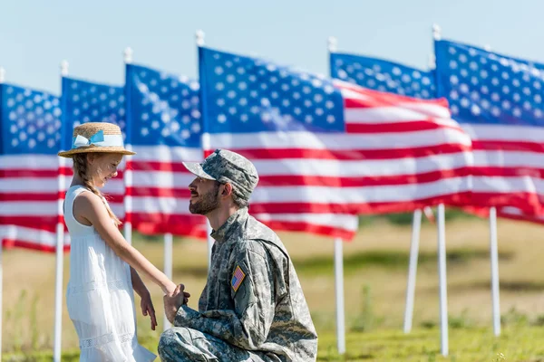 Glücklicher Militärmann, der ein Kind mit Strohhut ansieht, während er Händchen hält in der Nähe amerikanischer Flaggen mit Sternen und Streifen — Stockfoto