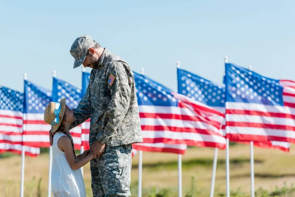 Щасливий військовий чоловік дивиться на милу дитину в солом'яному капелюсі біля американських прапорів з зірками і смугами — стокове фото