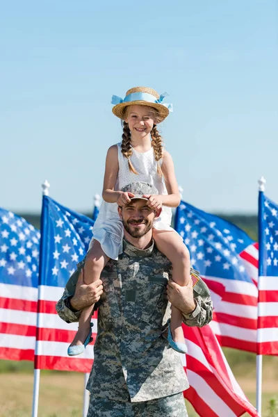 Schöner Mann hält auf Schultern glückliche Tochter mit Strohhut in der Nähe amerikanischer Flaggen — Stockfoto