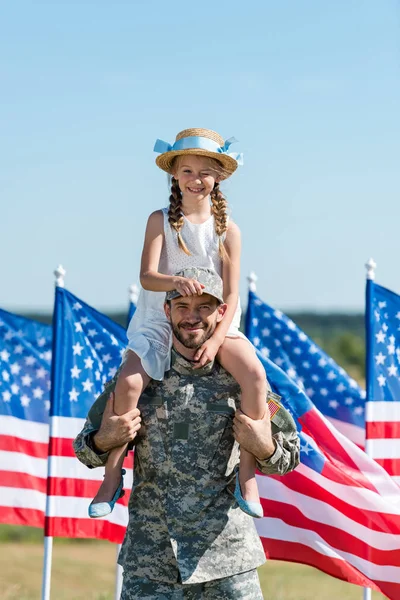 Schöner Mann hält auf Schultern fröhliche Tochter mit Strohhut in der Nähe amerikanischer Flaggen — Stockfoto