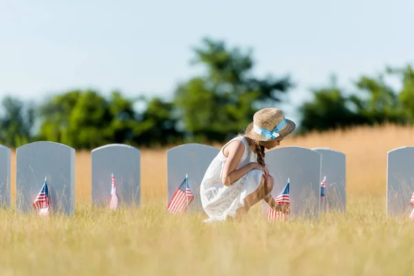 Selective focus of kid in dress and straw hat  sitting near headstone with american flag in graveyard — Stock Photo