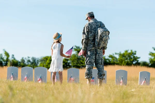 Criança de chapéu de palha e homem de uniforme militar em pé perto de lápides e segurando bandeiras americanas no cemitério — Fotografia de Stock