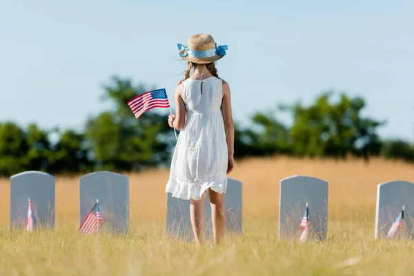 Rückansicht eines Kindes in Kleid und Strohhut, das neben Grabstein steht und die amerikanische Flagge auf dem Friedhof hält — Stockfoto