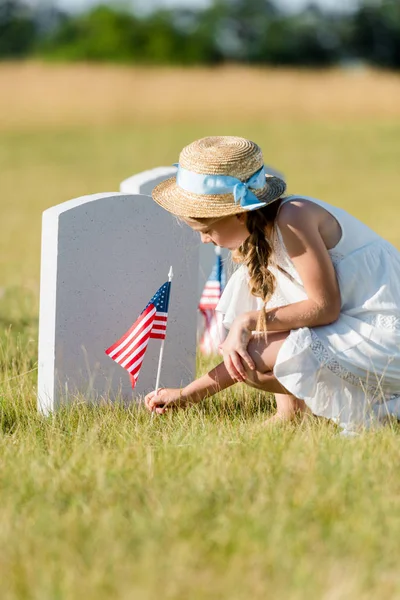 Selective focus of adorable kid sitting near headstone with american flag in graveyard — Stock Photo