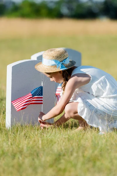 Selektiver Fokus eines entzückenden Kindes, das in der Nähe eines Grabsteins mit amerikanischer Flagge auf dem Friedhof sitzt — Stockfoto