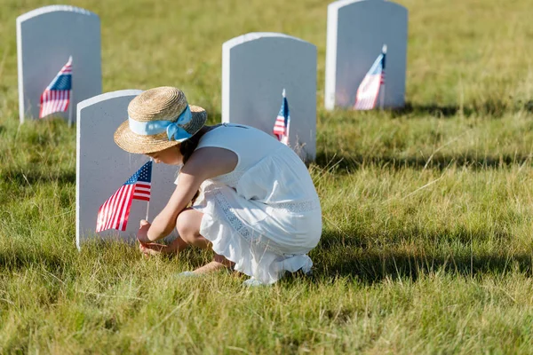 Selective focus of  kid in straw hat sitting near headstone with american flag in graveyard — Stock Photo