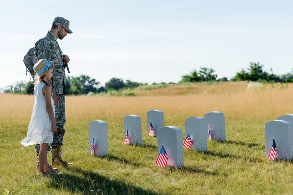 Soldat mit Mütze und Kind mit Strohhut betrachten Grabsteine auf Friedhof — Stockfoto