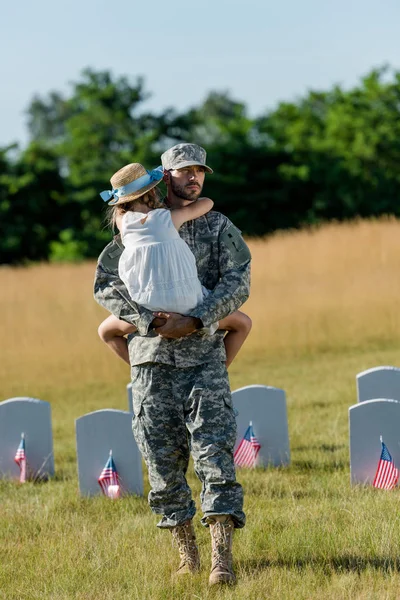 Militärmann mit Mütze hält ein Kind mit Strohhut in der Nähe von Grabsteinen mit amerikanischen Flaggen — Stockfoto