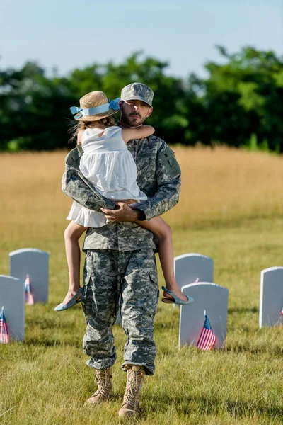 Père militaire en casquette tenant dans ams enfant en chapeau de paille près des pierres tombales avec des drapeaux américains — Photo de stock