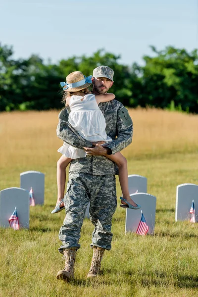 Ветеранский отец в кепке держа в руках ребенка в соломенной шляпе рядом с надгробиями с американскими флагами — стоковое фото
