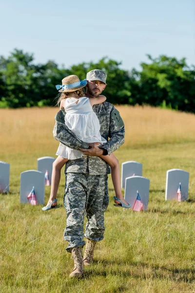 Veterano em cap segurando em ams criança em chapéu de palha perto de lápides com bandeiras americanas — Fotografia de Stock