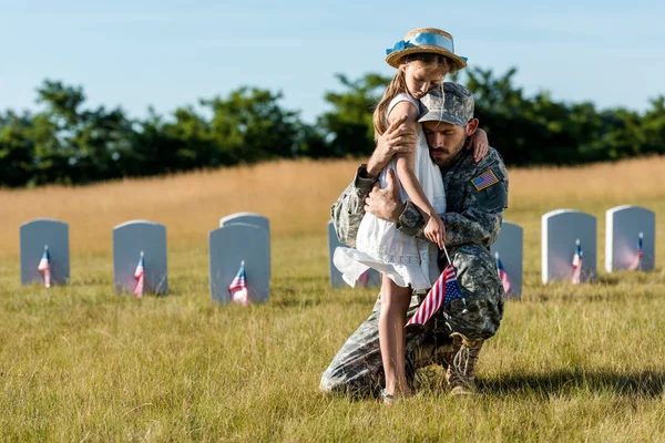 Militar en uniforme abrazando hija cerca de lápidas en cementerio - foto de stock
