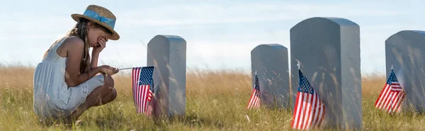 Panoramic shot of kid in straw hat covering face while sitting near headstones with american flags — Stock Photo