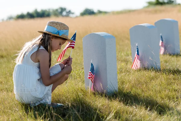 Triste enfant en chapeau de paille assis près des pierres tombales et tenant drapeau américain — Photo de stock