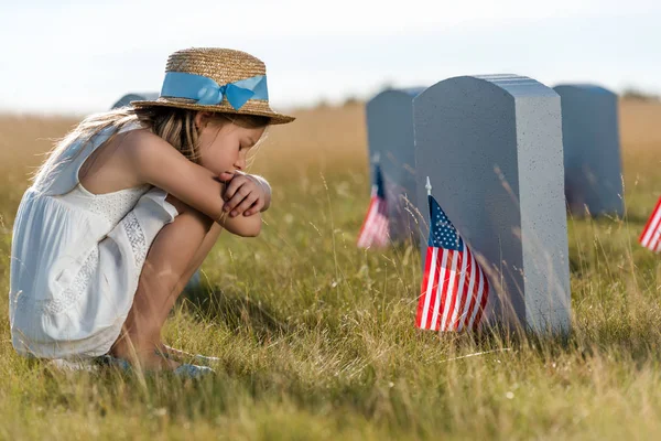 Triste enfant en chapeau de paille assis près des pierres tombales avec des drapeaux américains — Photo de stock